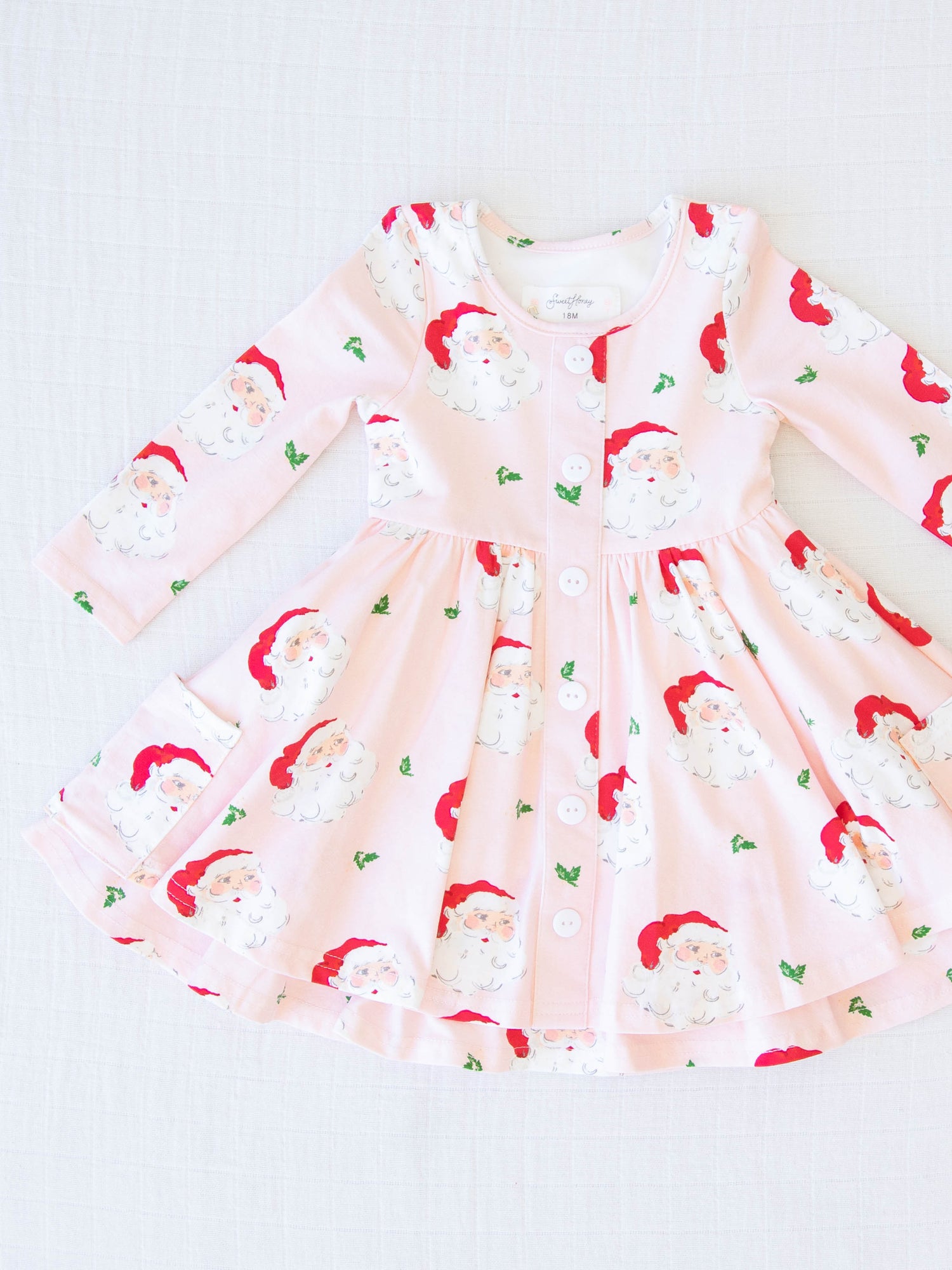 Prim Dress - Dear Santa. This long-sleeved dress features a pattern of large Santa Claus heads on a pale pink background.