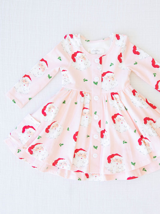 Prim Dress - Dear Santa. This long-sleeved dress features a pattern of large Santa Claus heads on a pale pink background.