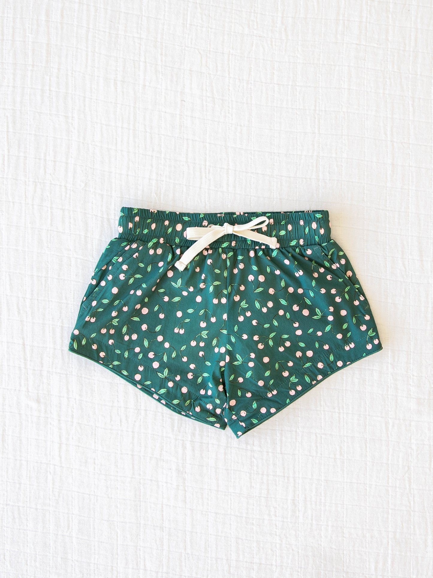 European cut style of our Boy's Everyday Lined Trunks – Cherry on Top. This pair of trunks is a pattern of light pink cherries on a dark green background.