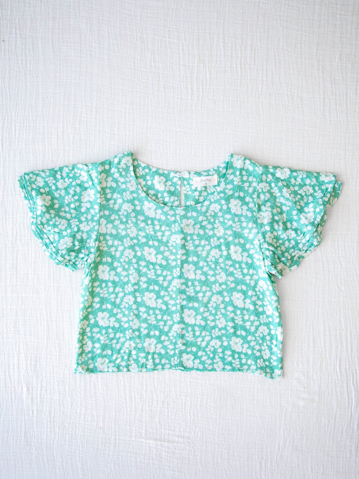 Classic Flutter Top - Green Rush. This top has a keyhole back and flowy double ruffle sleeves. It is a pattern of white flowers and leaves on a vibrant green background.