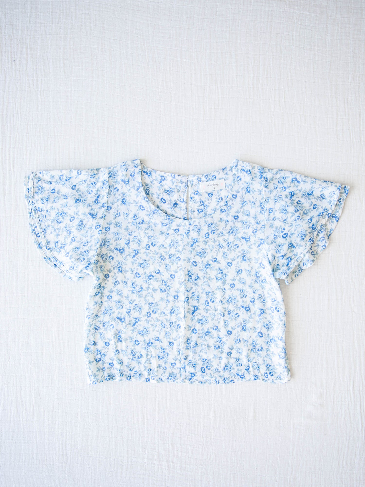 Classic Flutter Top - Blue Jay Floral. This top has a keyhole back and flowy double ruffle sleeves. It is a blue rose pattern on a pale background.