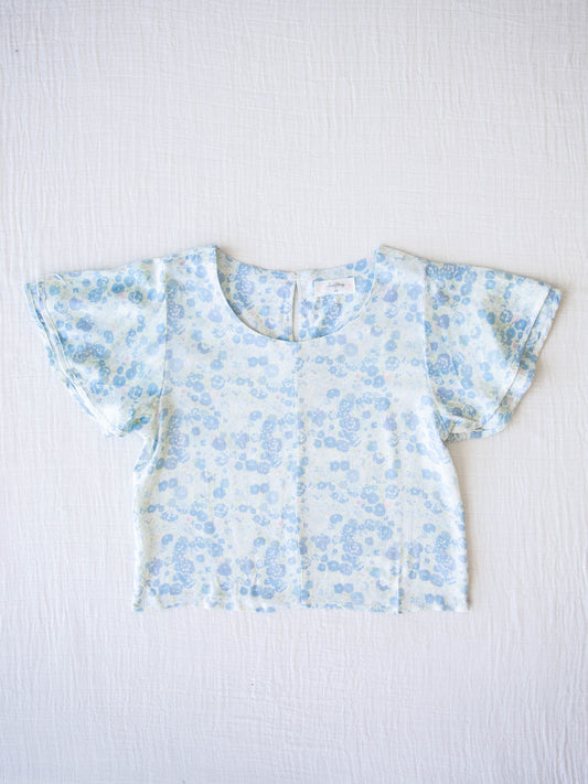 Classic Flutter Top - Pastel Floral. This top has a keyhole back and flowy double ruffle sleeves. It is a pattern of blue flowers and green stems on an ivory background.