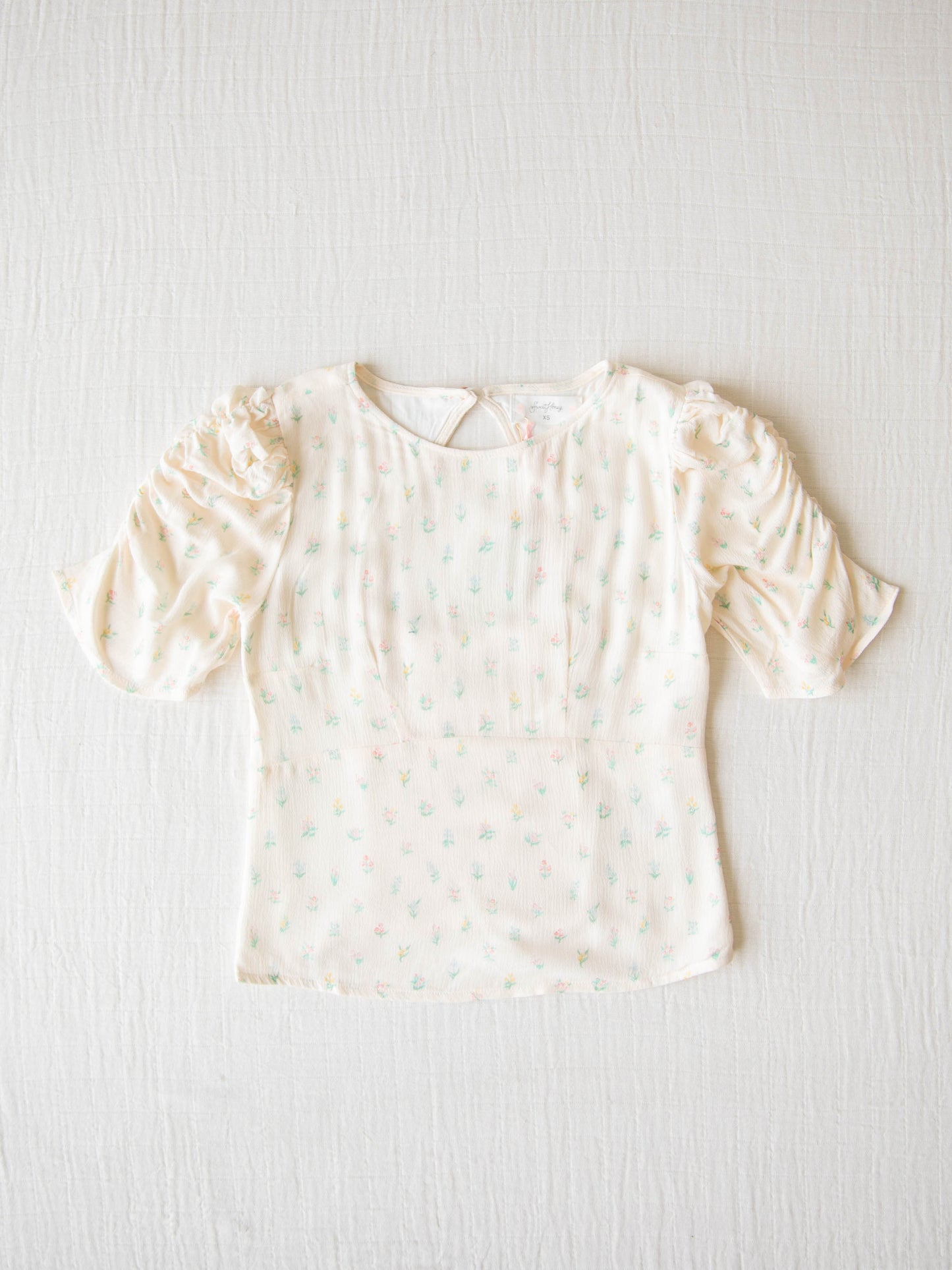 Classic Smocked Cutout Top – Floral Drop. This short sleeve top has a smocked and keyhole back, and synched sleeves. It is a pattern of tiny hand drawn flowers spaced out across a background of cream.