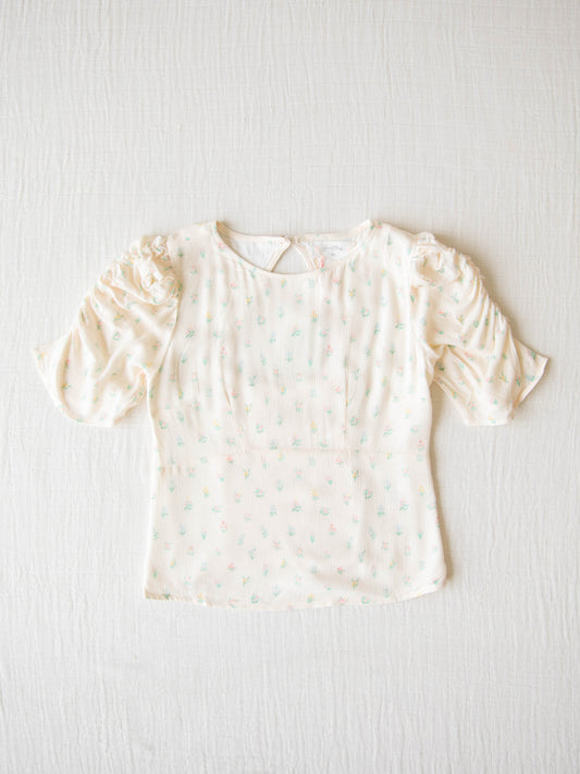 Classic Smocked Cutout Top – Floral Drop. This short sleeve top has a smocked and keyhole back, and synched sleeves. It is a pattern of tiny hand drawn flowers spaced out across a background of cream.