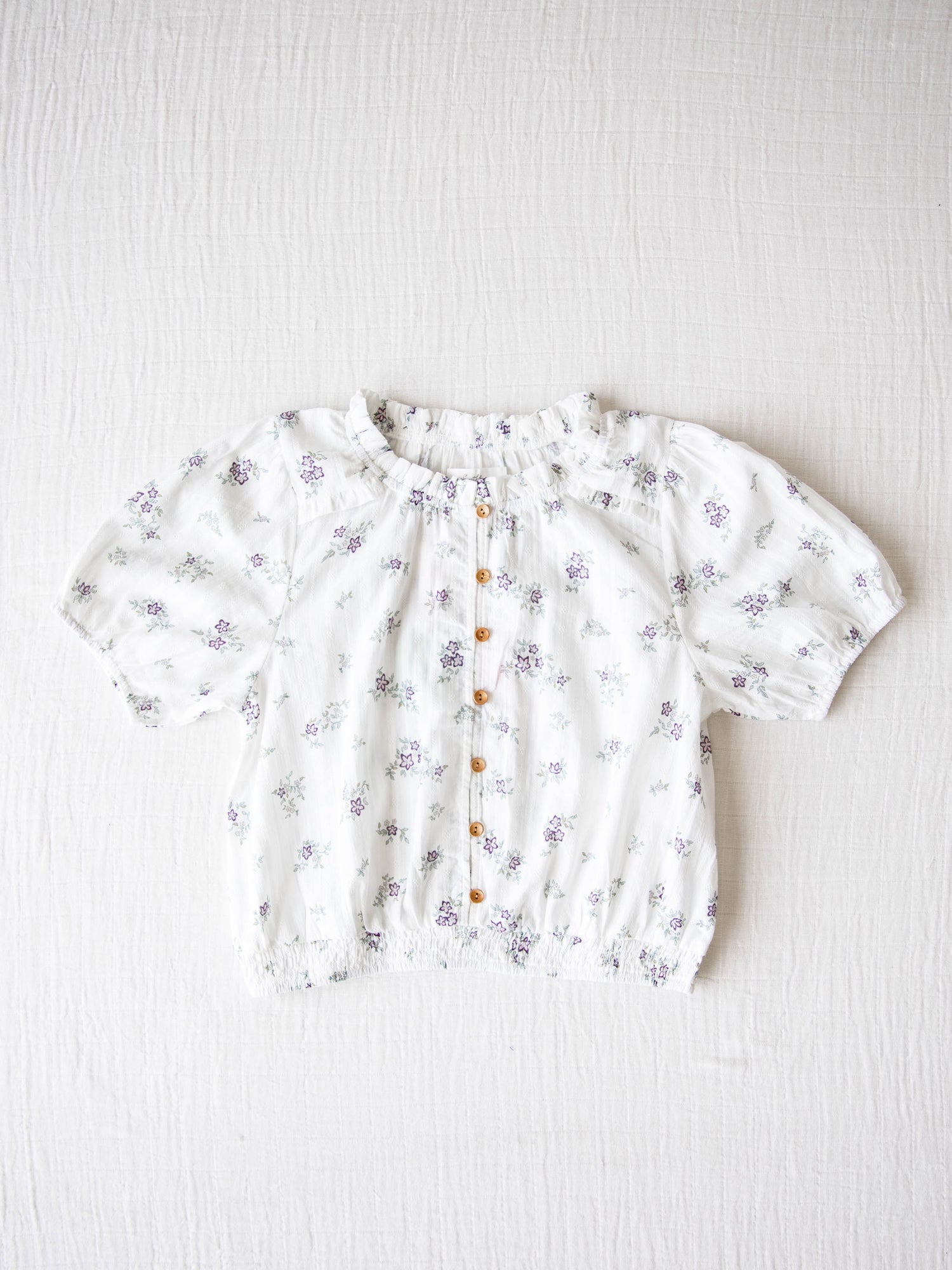 Classic Button Top - Lavender Floral. This top has functional buttons down the bodice, puff sleeves, and elastic ruffle neckline. It is a pattern of lavender and purple colored blooms with leaves on an ivory background.