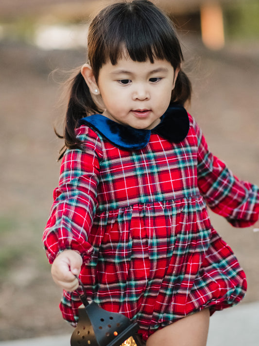 This image of a girl features the product Classic Bubble - Noel Plaid. This Classic Bubble has a peter pan collar and buttons down the back. It is red, green, black, and white plaid.