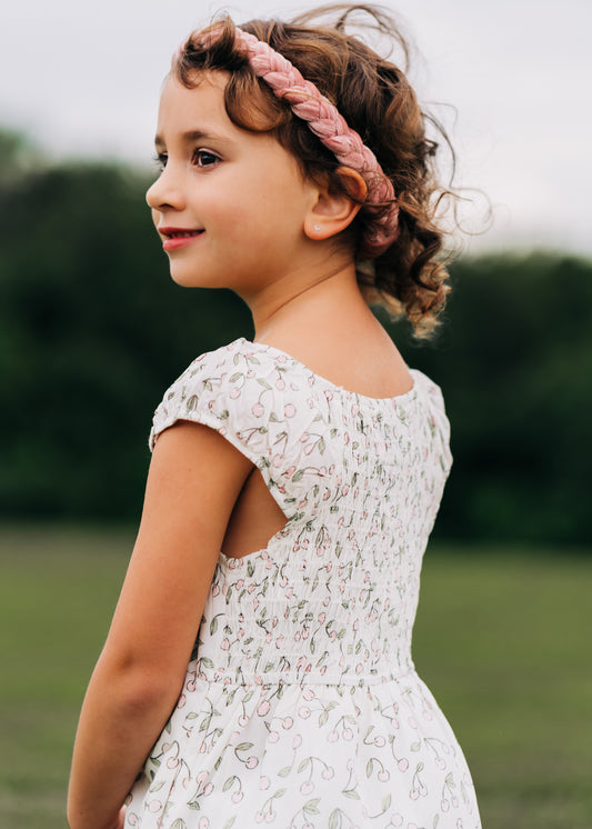 This image of a girl features the product Smocked Dress – Cherry in Pink. This short dress comes in a pattern of pink cherries with green leaves over an ivory background.