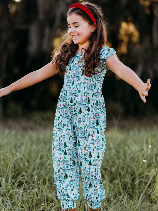 This image of a girl features the product Smocked Romper - Christmas Town. This short sleeved smocked romper comes in a pattern of Christmas themed buildings and evergreens on a snowy background.
