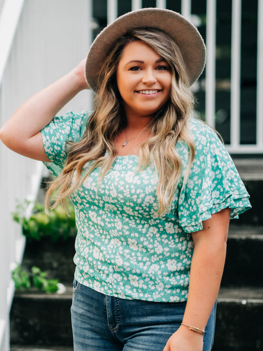 This image of a woman features the product Classic Flutter Top - Green Rush. This top has a keyhole back and flowy double ruffle sleeves. It is a pattern of white flowers and leaves on a vibrant green background.