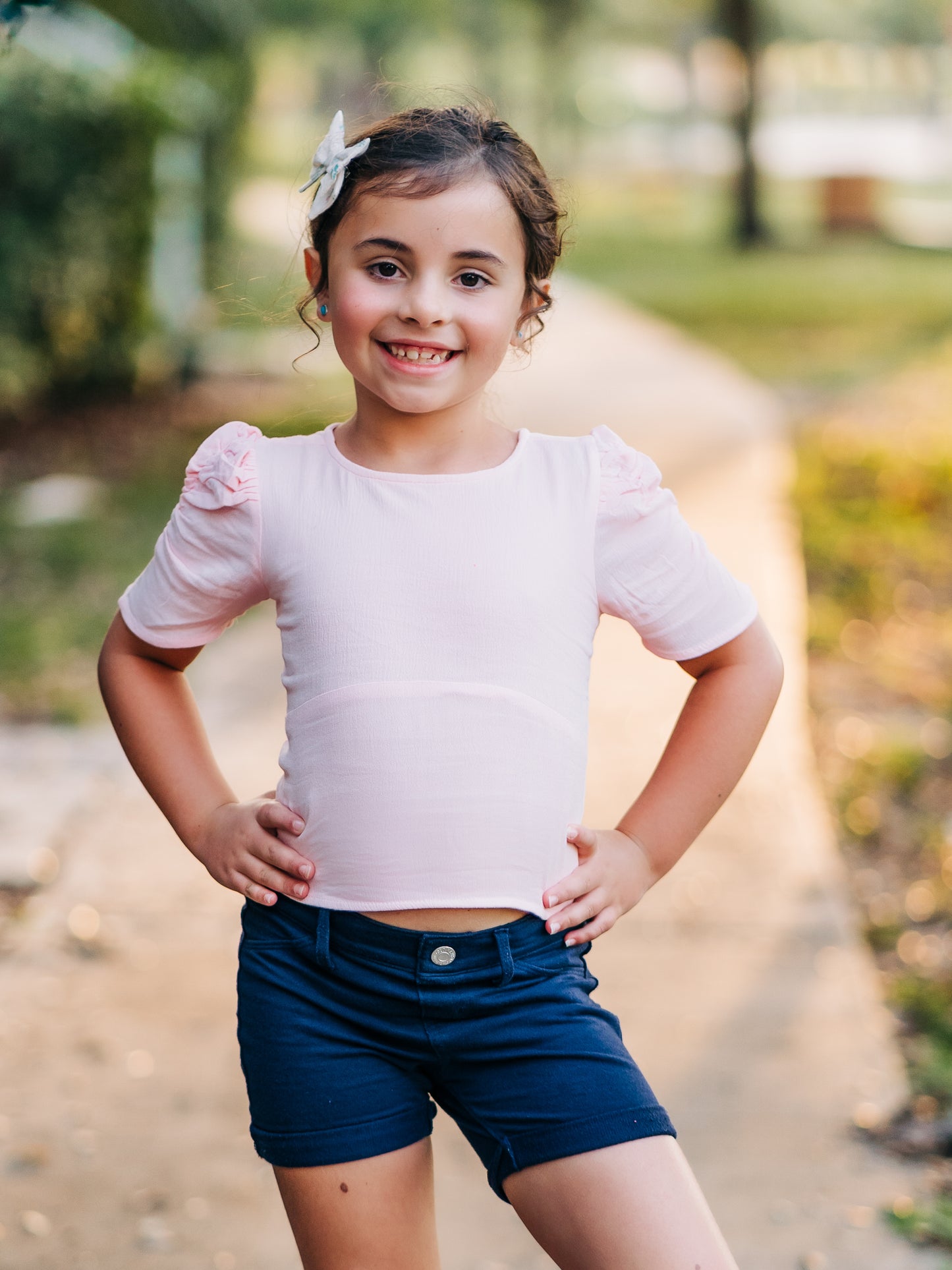 This image of a girl features the product Classic Smocked Cutout Top – Soft Pink. This short sleeve top has a smocked and keyhole back, and synched sleeves. 
