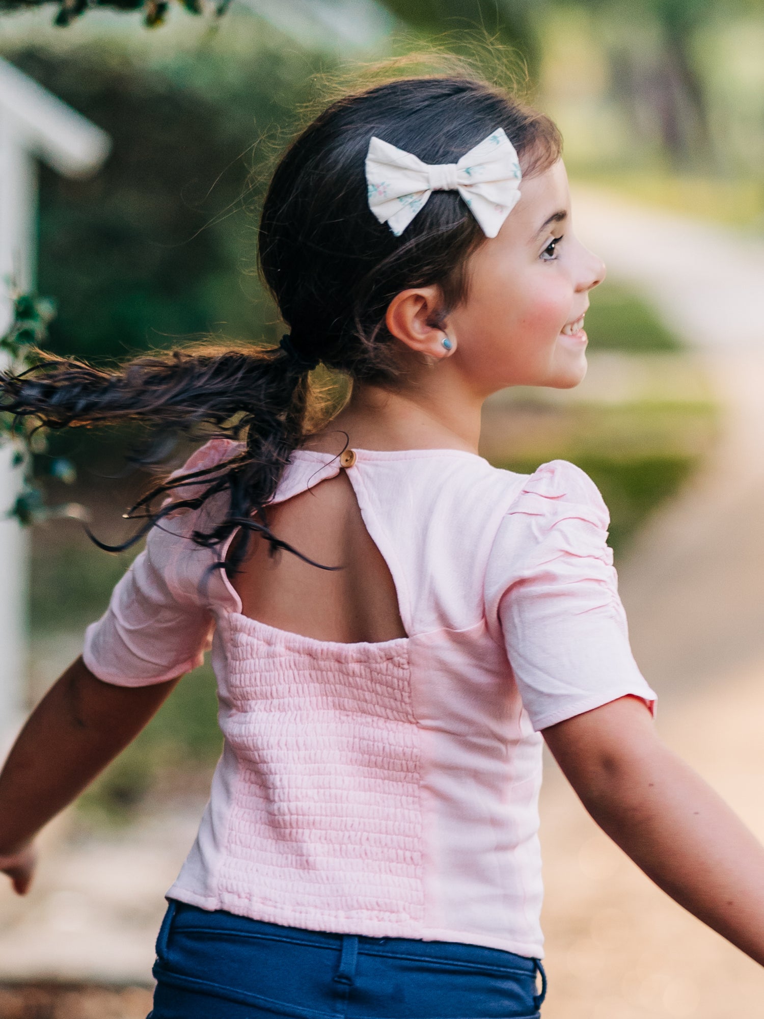 This image of a girl features the product Classic Smocked Cutout Top – Soft Pink. This short sleeve top has a smocked and keyhole back, and synched sleeves. 