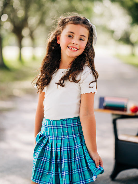 This image of a girl features the product Pleated Skort – Whimsical Plaid. This pleated skirt comes with built-in shorts and an elastic back in a teal, dark blue, and white plaid.