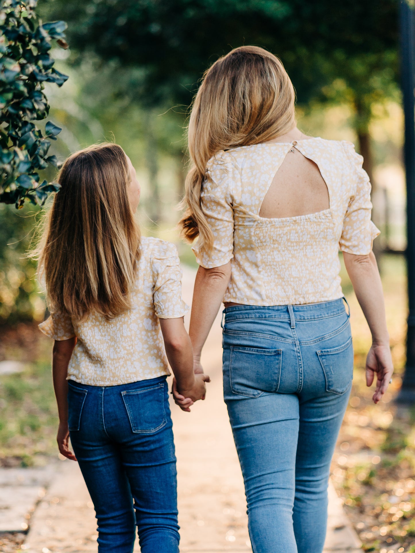 This image of a woman and her daughter features the product Classic Smocked Cutout Top – I’ve Got Sunshine. This short sleeve top has a smocked and keyhole back, and synched sleeves. It is a pattern of white flowers on a bright yellow background.