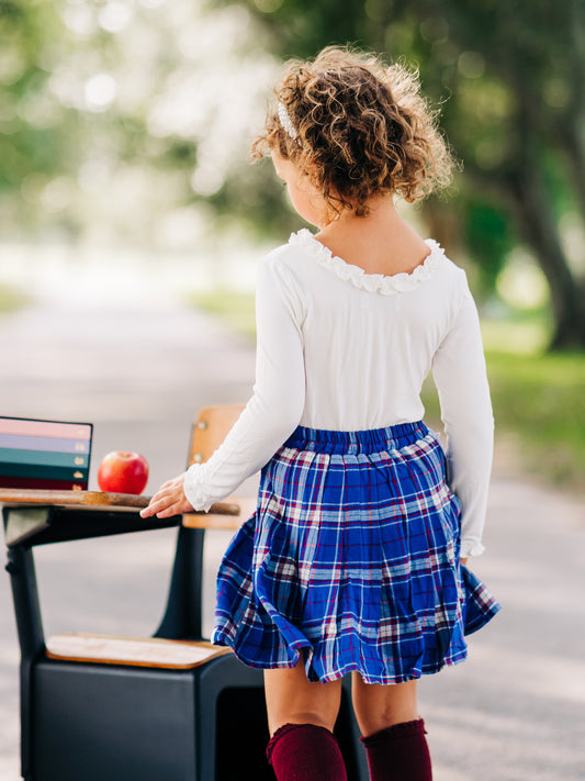 This image of a girl features the product Pleated Skort – Royal Blue Check. This pleated skirt comes with built-in shorts and an elastic back in a plaid pattern of royal blue, red and white.