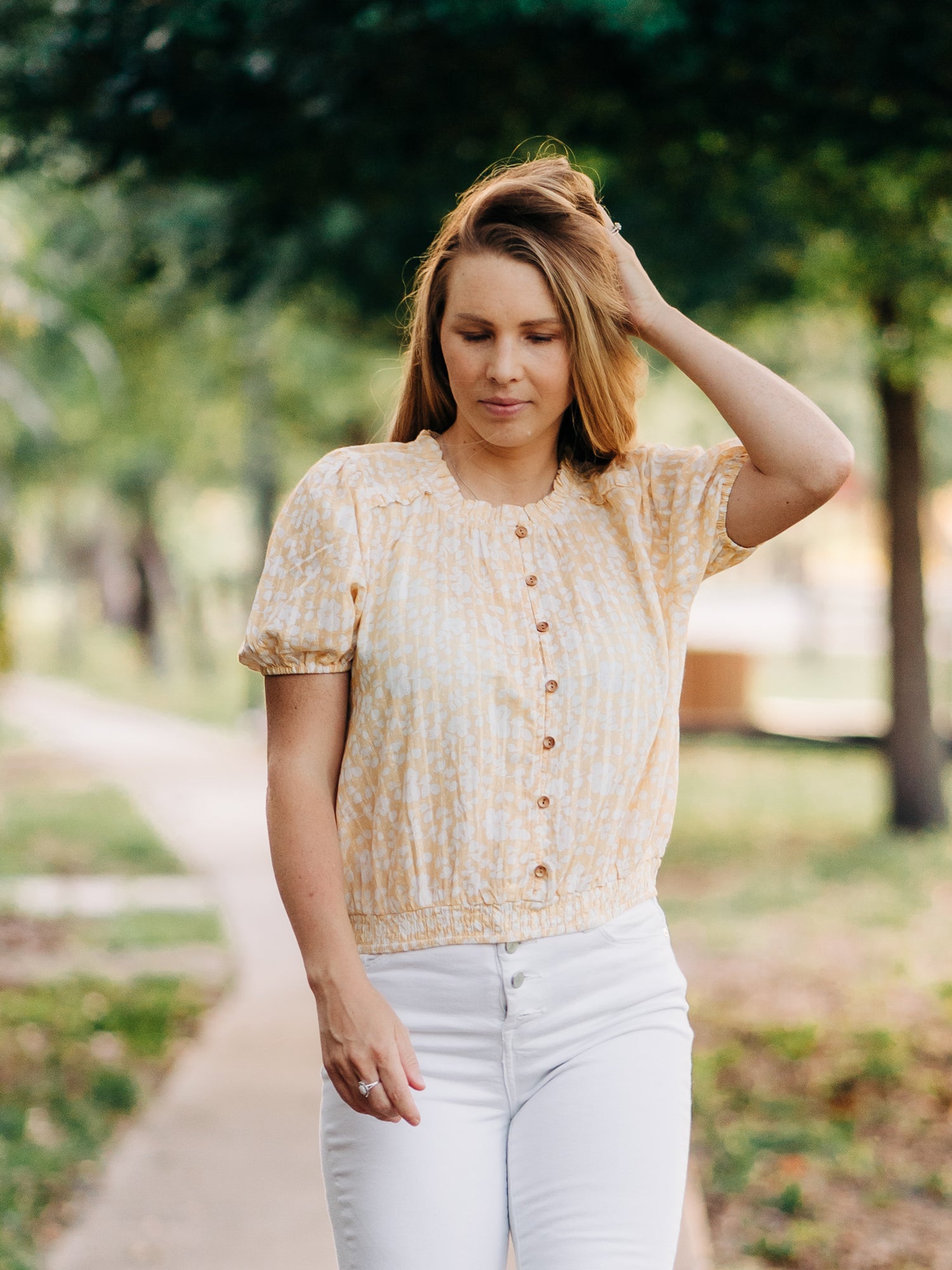 This image of a woman features the product Classic Button Top - I’ve Got Sunshine. This top has functional buttons down the bodice, puff sleeves, and elastic ruffle neckline. It is a pattern of white flowers on a bright yellow background.