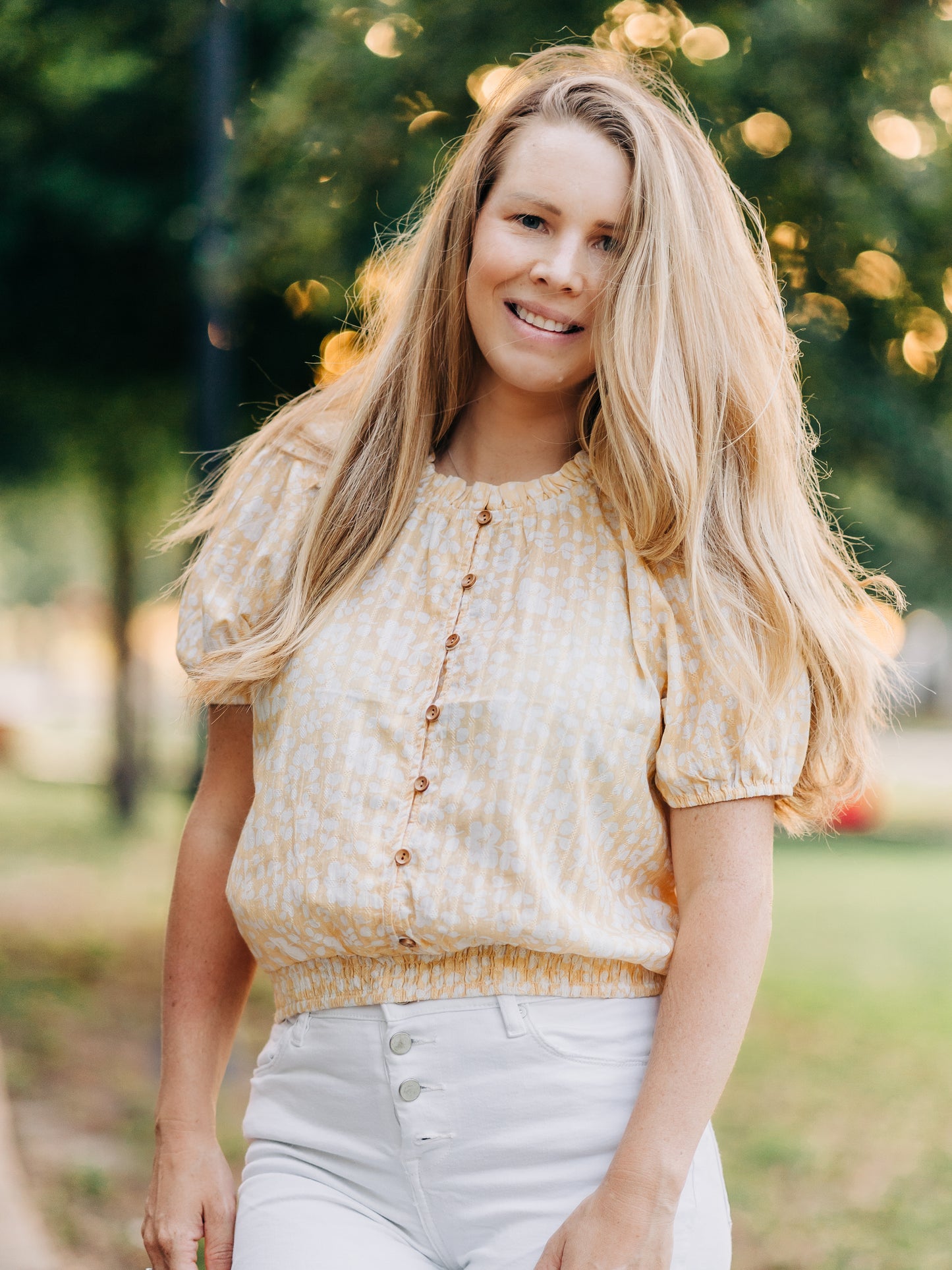 This image of a woman features the product Classic Button Top - I’ve Got Sunshine. This top has functional buttons down the bodice, puff sleeves, and elastic ruffle neckline. It is a pattern of white flowers on a bright yellow background.
