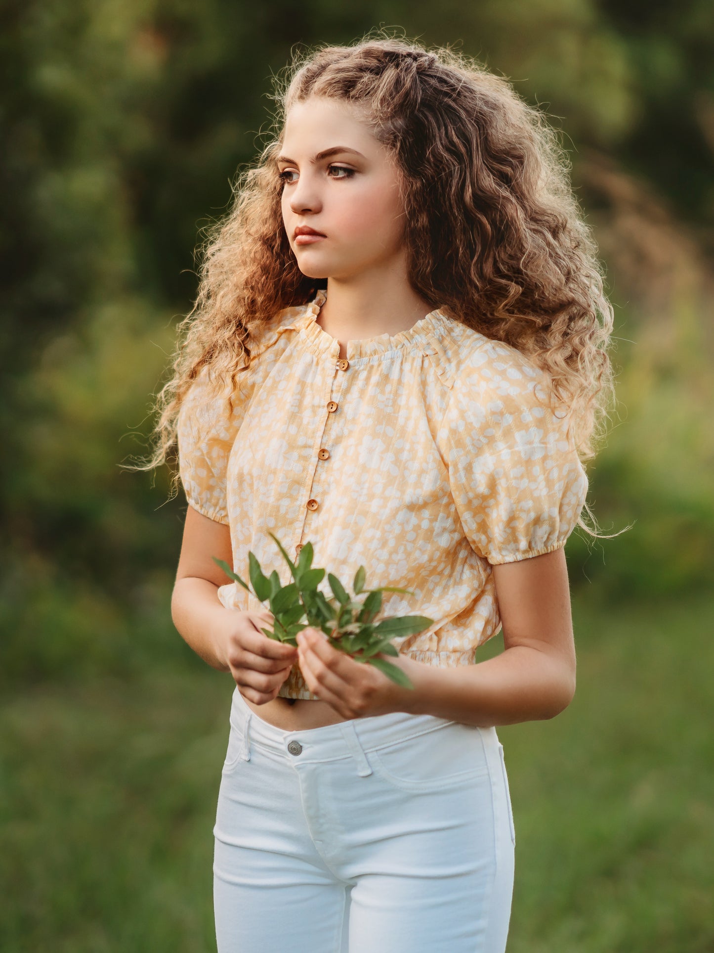 This image of a girl features the product Classic Button Top - I’ve Got Sunshine. This top has functional buttons down the bodice, puff sleeves, and elastic ruffle neckline. It is a pattern of white flowers on a bright yellow background.