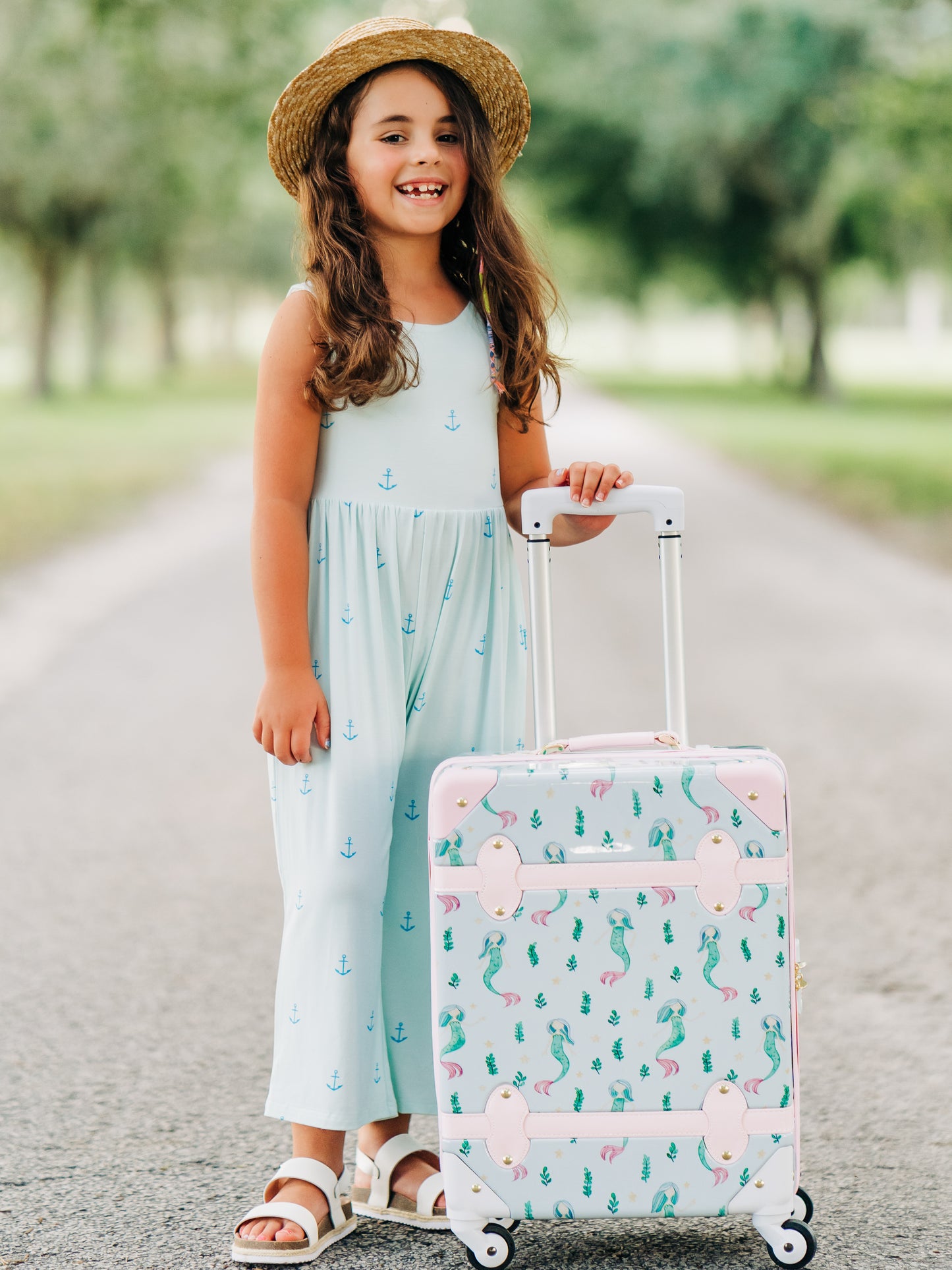 This image of a girl features the product Lennon Traveling Luggage – Sea Princess. The outside of this luggage is a pattern of mermaids and little green sea plants on a pale blue background. The inside lining is a pattern of pale blue starfish like stars on a blue background.