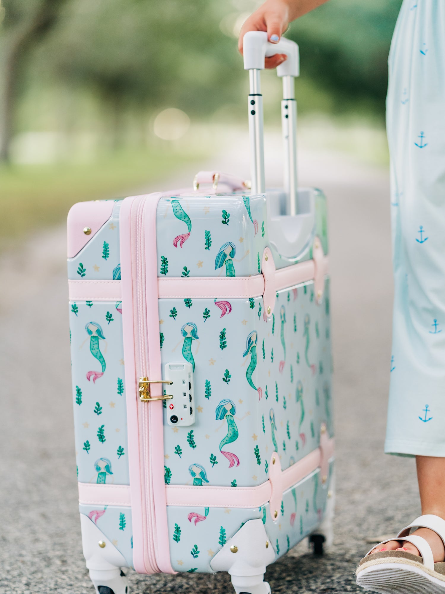 This image of a girl features the product Lennon Traveling Luggage – Sea Princess. The outside of this luggage is a pattern of mermaids and little green sea plants on a pale blue background. The inside lining is a pattern of pale blue starfish like stars on a blue background.