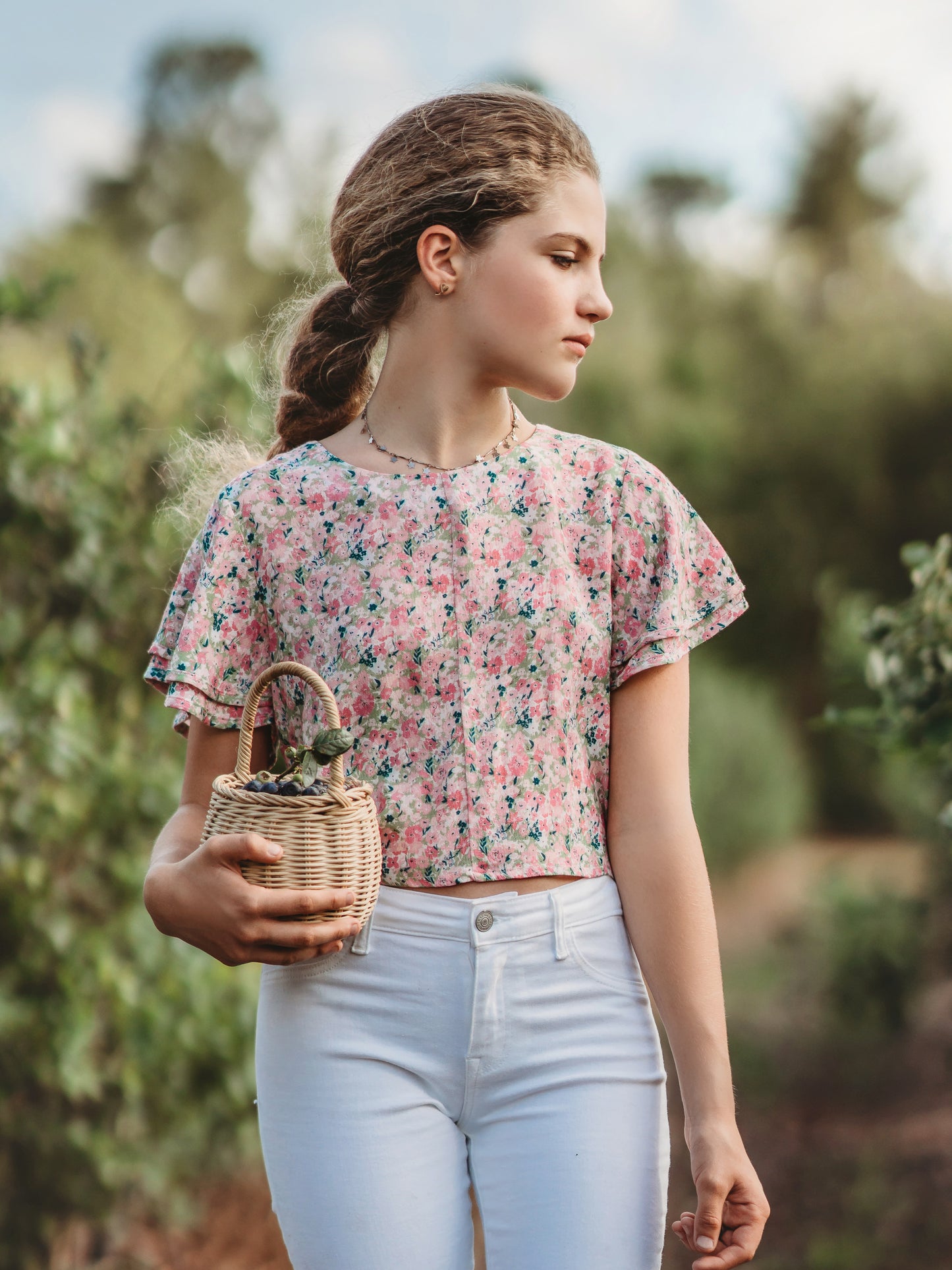 This image of a young woman features the product Classic Flutter Top - Joyful. This top has a keyhole back and flowy double ruffle sleeves. It is a pink and green floral pattern.