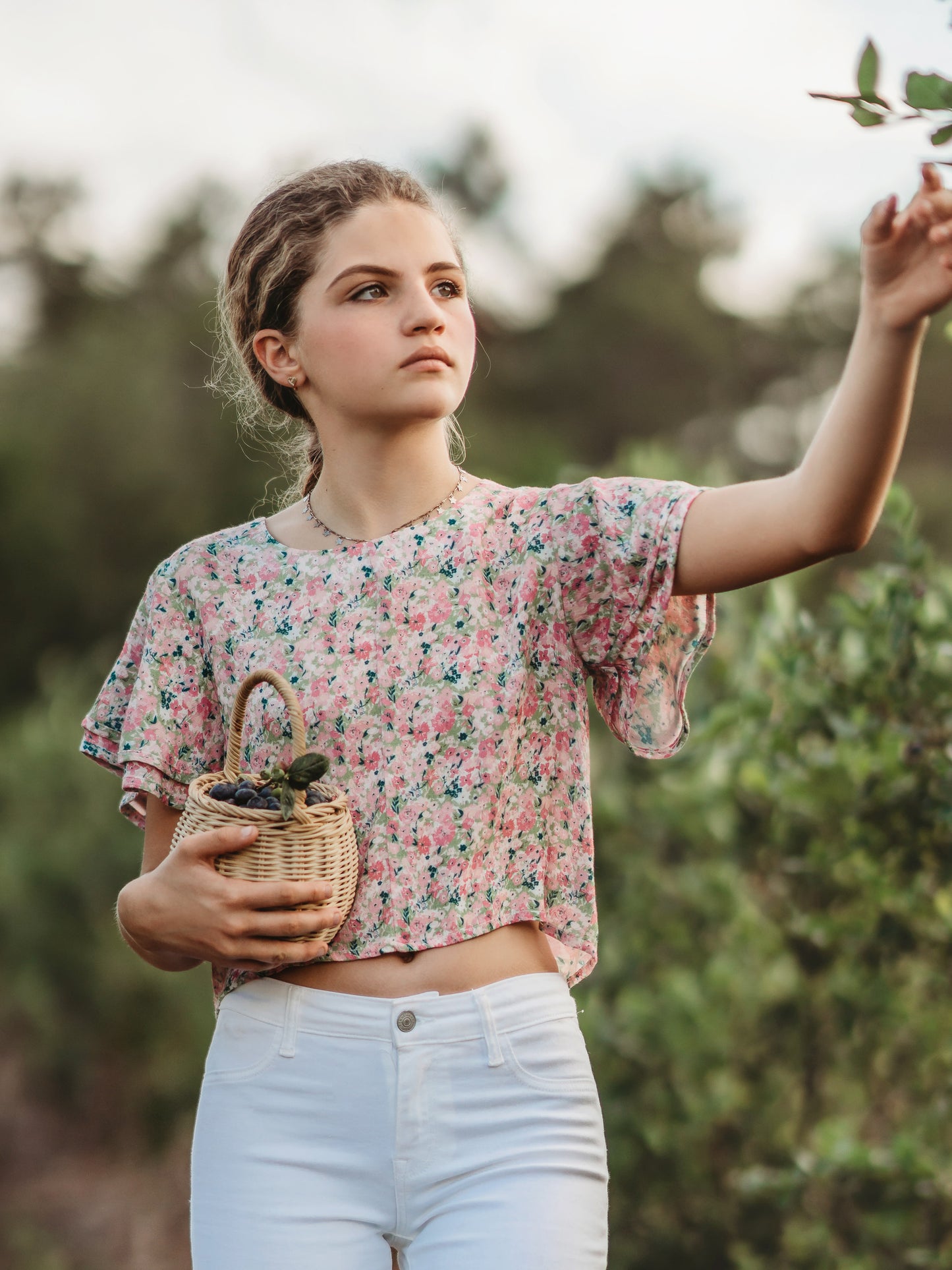 This image of a young woman features the product Classic Flutter Top - Joyful. This top has a keyhole back and flowy double ruffle sleeves. It is a pink and green floral pattern.