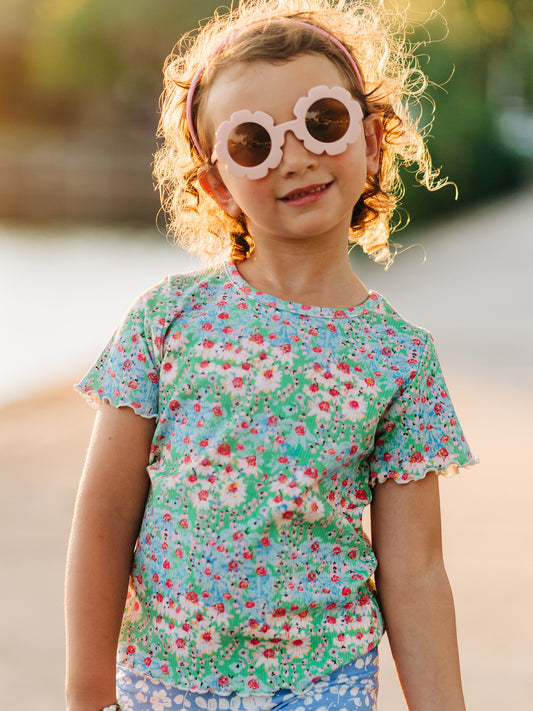 This image of a girl features the product Frilled Sleeve Shirt - Wildflowers. It comes in a pattern of light blue and pink flowers on a green background.