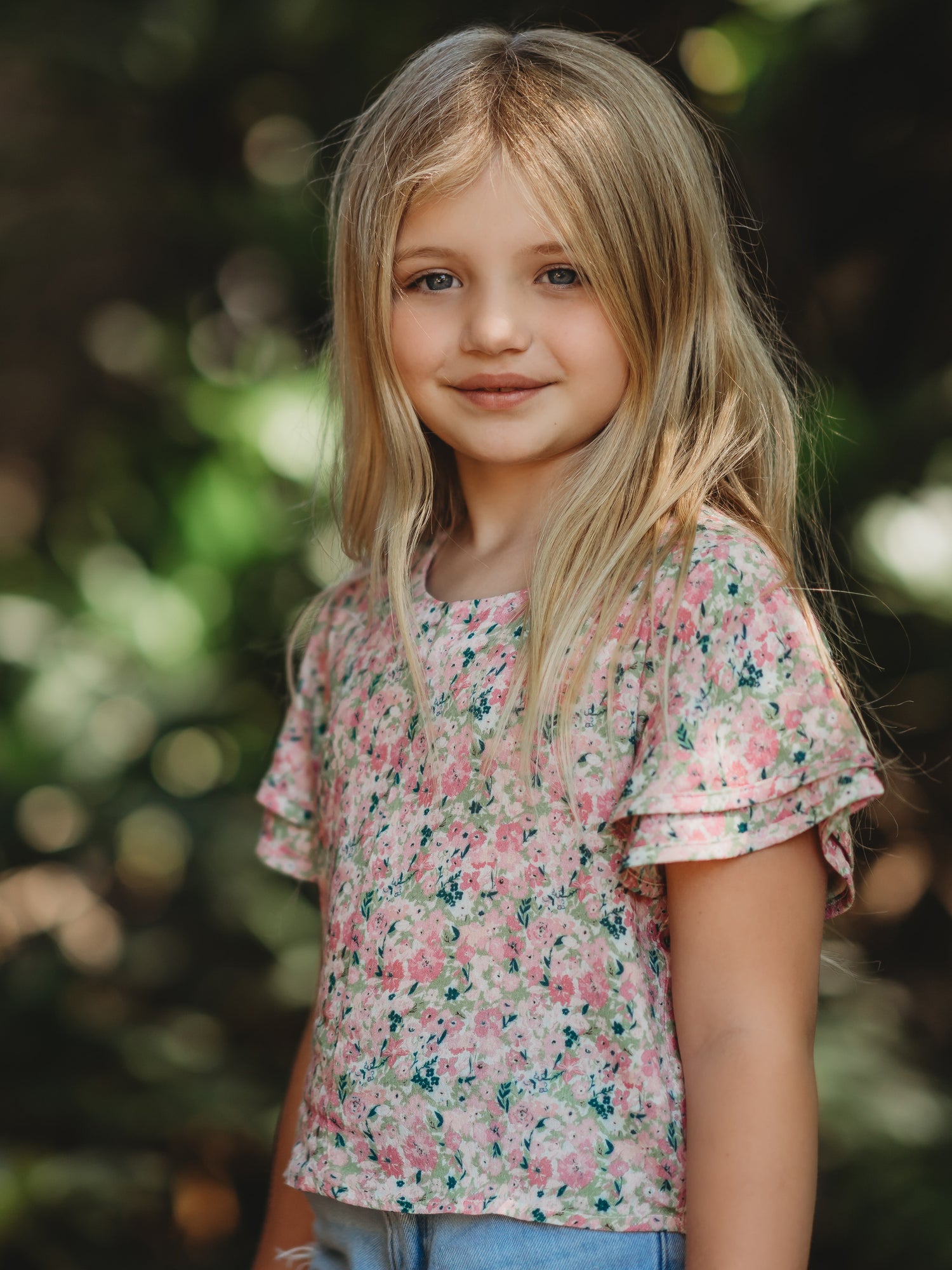 This image of a girl features the product Classic Flutter Top - Joyful. This top has a keyhole back and flowy double ruffle sleeves. It is a pink and green floral pattern.