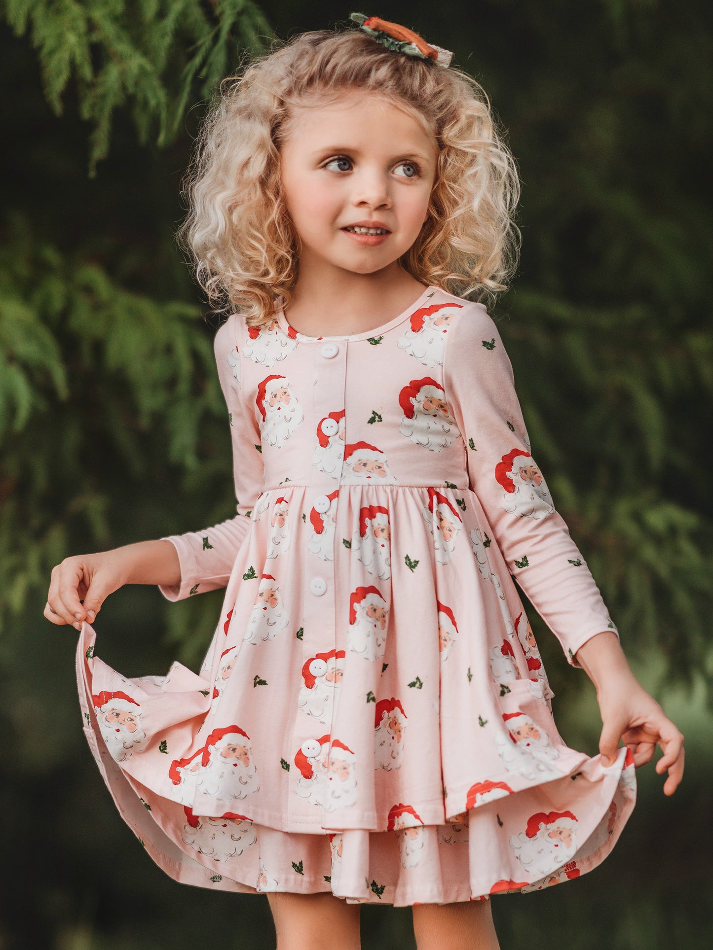 This image of a girl features the product Prim Dress - Dear Santa. This long-sleeved dress features a pattern of large Santa Claus heads on a pale pink background.