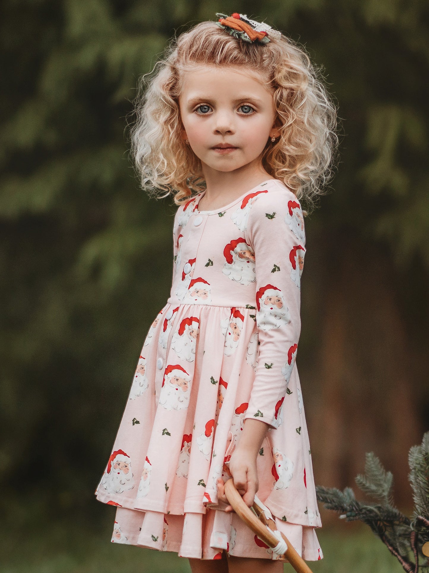 This image of a girl features the product Prim Dress - Dear Santa. This long-sleeved dress features a pattern of large Santa Claus heads on a pale pink background.