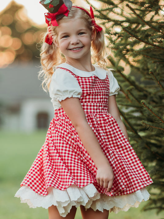 This image of a girl features the product Pinafore - Peppermint Check. This crisscross strapped pinafore comes in a red and white check pattern.