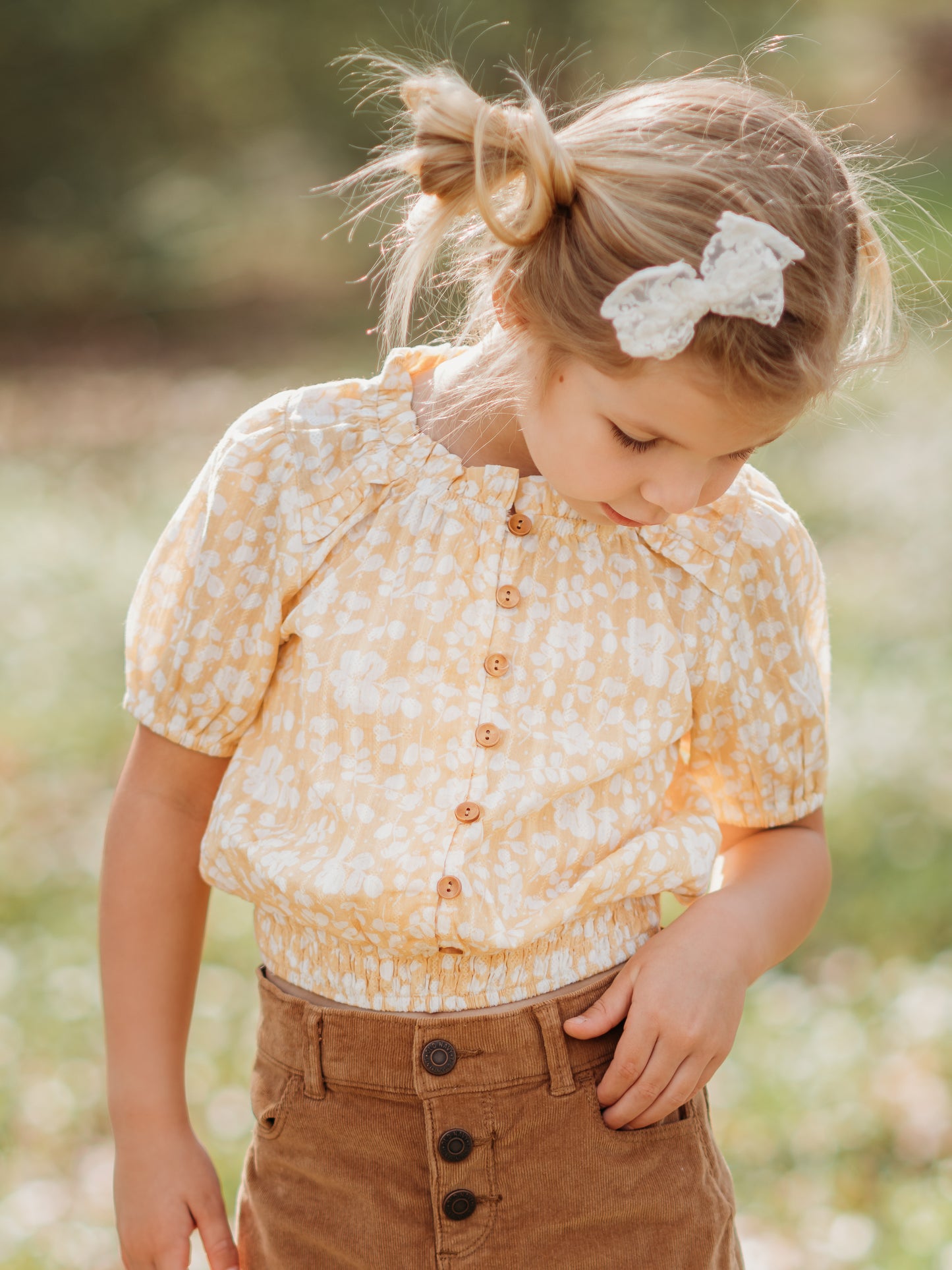 This image of a girl features the product Classic Button Top - I’ve Got Sunshine. This top has functional buttons down the bodice, puff sleeves, and elastic ruffle neckline. It is a pattern of white flowers on a bright yellow background.