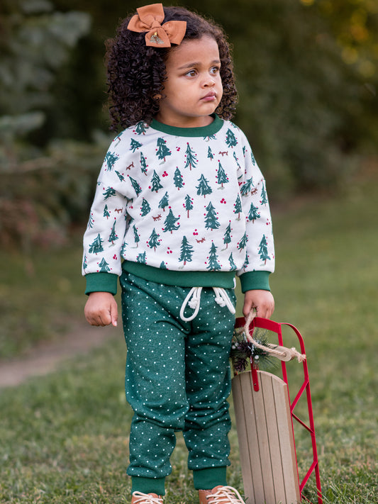 This image of a girl features the product Sweatshirt Set - O Christmas Tree. The top is a pattern of evergreen trees with red ornaments on a snowy background. The bottoms come in a pattern of white polka dots on a deep green background.