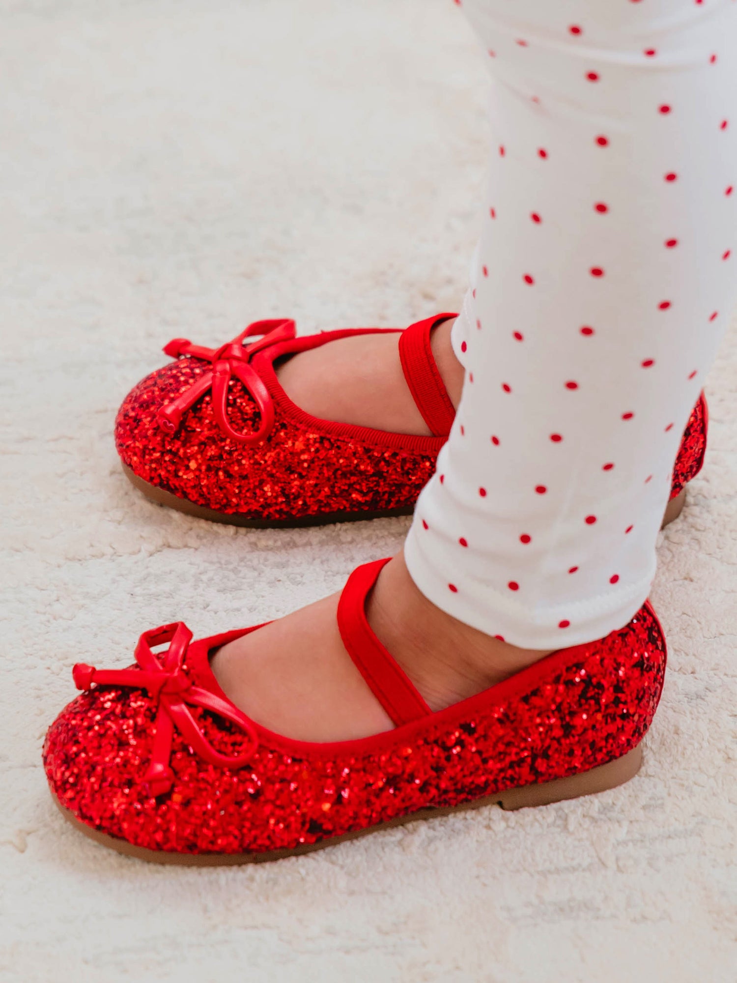 These Small Bow Glitter Ballet shoes come in red with an elastic strap across the top of the foot. The red bows bows are attached on top of the shoes’ toe.