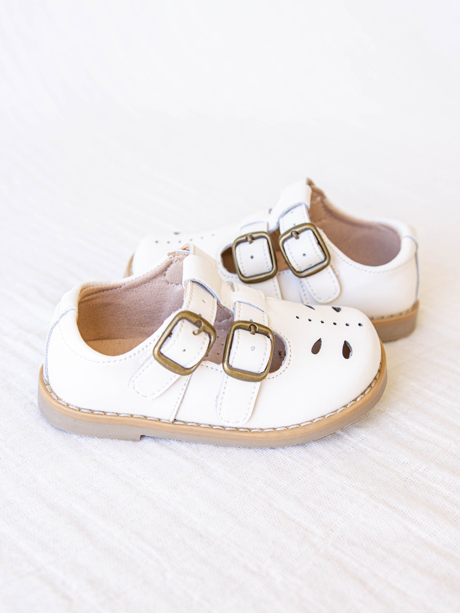 A pair of dove (white) colored genuine leather rubber soled shoes with two adjustable metal buckles and cut out detail across the toe area in a dragonfly type pattern. 