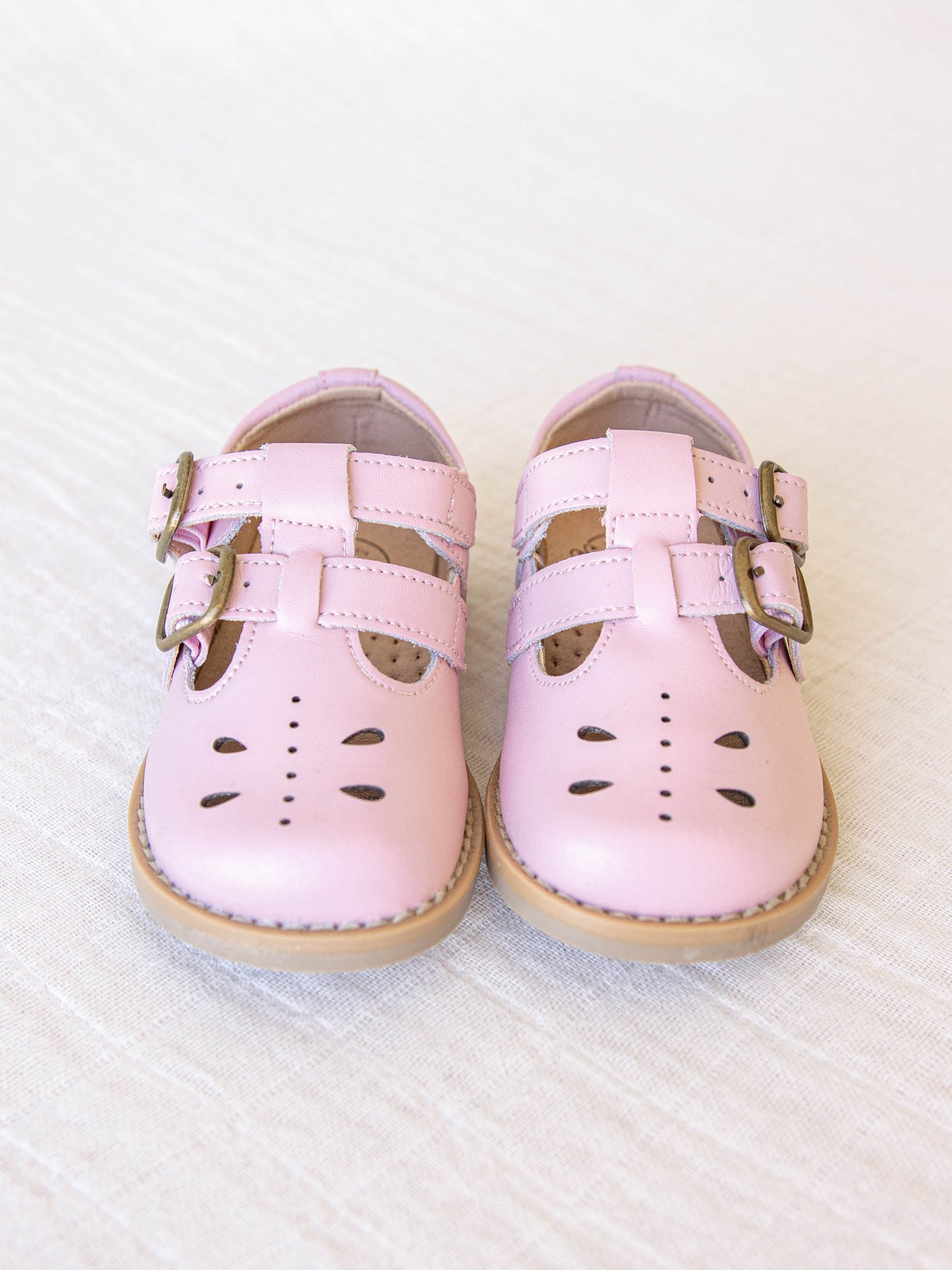 A pair of light pink colored genuine leather rubber soled shoes with two adjustable metal buckles and cut out detail across the toe area in a dragonfly type pattern. 
