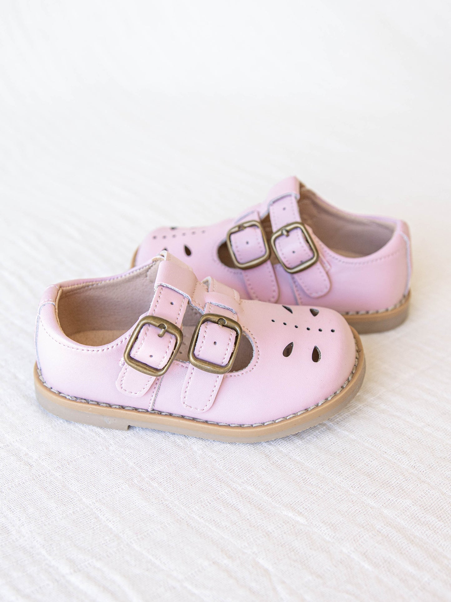 A pair of light pink colored genuine leather rubber soled shoes with two adjustable metal buckles and cut out detail across the toe area in a dragonfly type pattern. 
