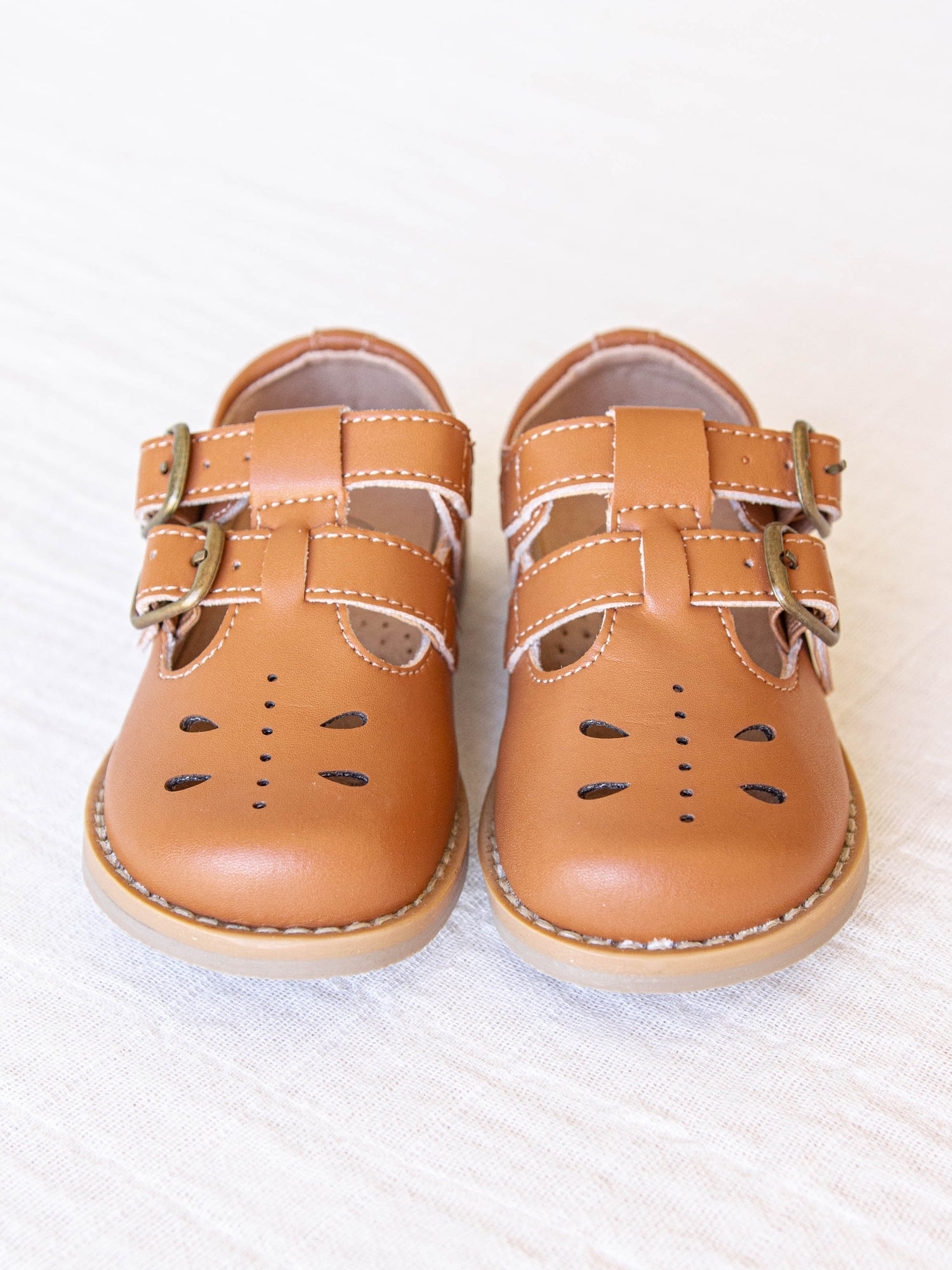 A pair of camel colored genuine leather rubber soled shoes with two adjustable metal buckles and ventilation holes across the toe area in a dragonfly type pattern. 