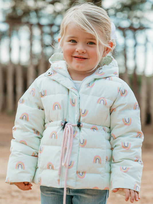 This image of a girl features the product Puffer Jacket - Over the Rainbow. It comes in a pattern of alternating pastel rainbows on a blue tinted background.