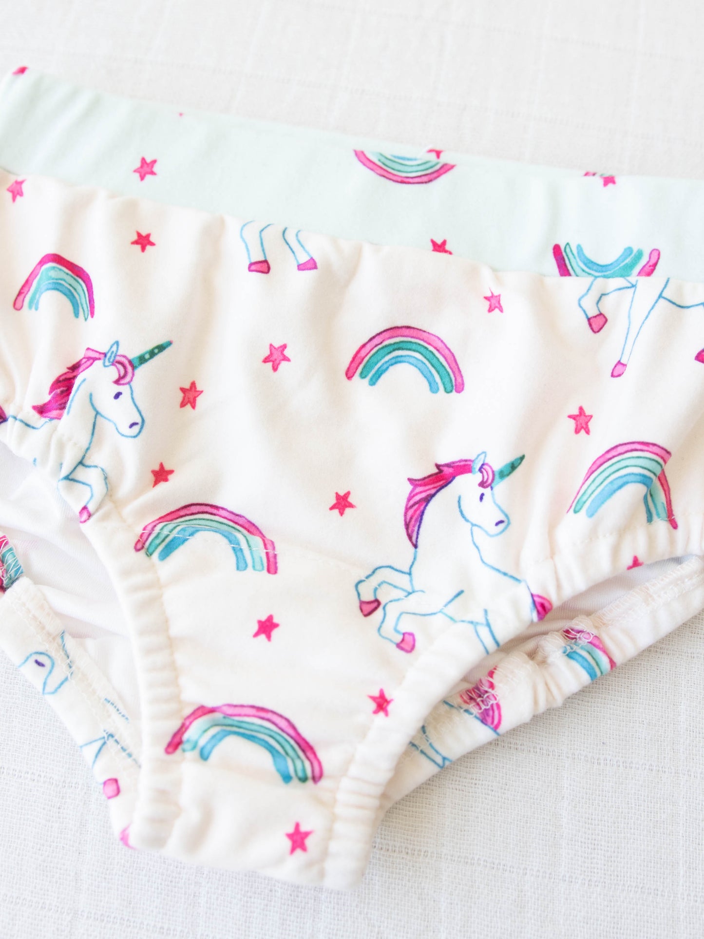 This pair of girls panties features the pattern Unicorn Dreams which has unicorns, rainbows, and stars over a pale pink background.