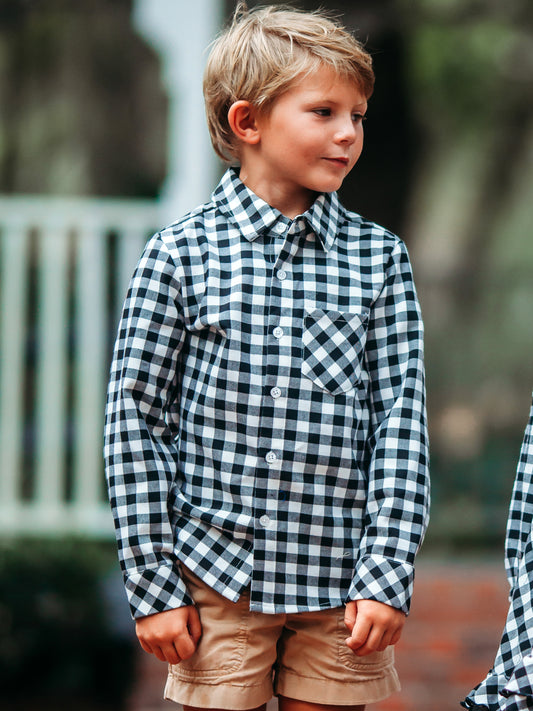 This image of a boy features the product Boy's Button Up Shirt - Black Plaid. This long sleeve button up shirt is a black, white, and grey check pattern.