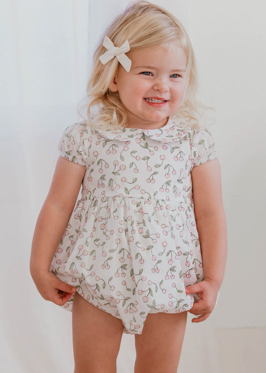 This image of a girl features the product Classic Bubble - Cherry in Pink. This Classic Bubble has a peter pan collar and buttons down the back. It is a pattern of pink cherries with green leaves over an ivory background.