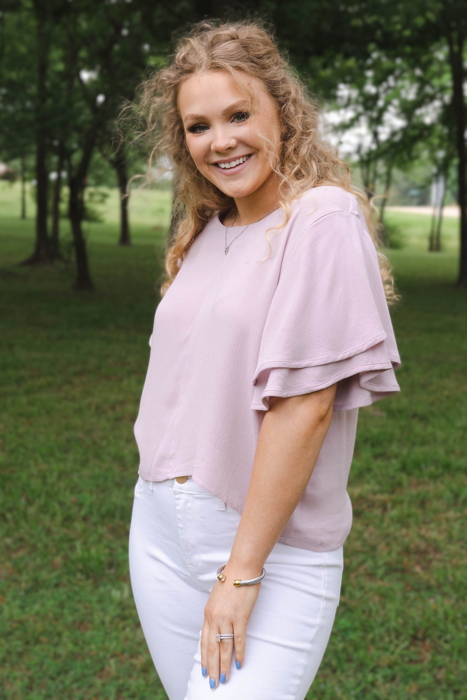 This image of a woman features the product Classic Flutter Top - Dusty Lavender. This top has a keyhole back and flowy double ruffle sleeves. It is a lovely dusty pink-purple color.