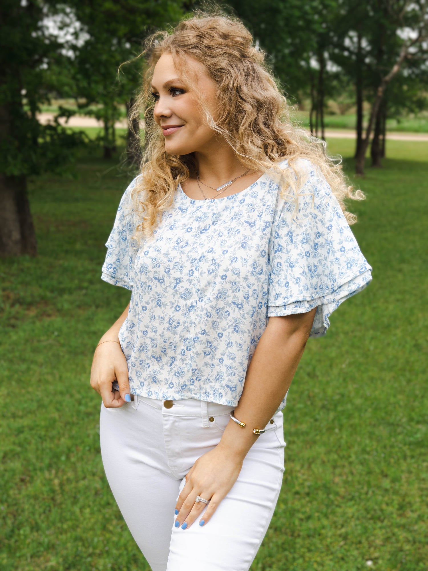 This image of a woman features the product Classic Flutter Top - Blue Jay Floral. This top has a keyhole back and flowy double ruffle sleeves. It is a blue rose pattern on a pale background.
