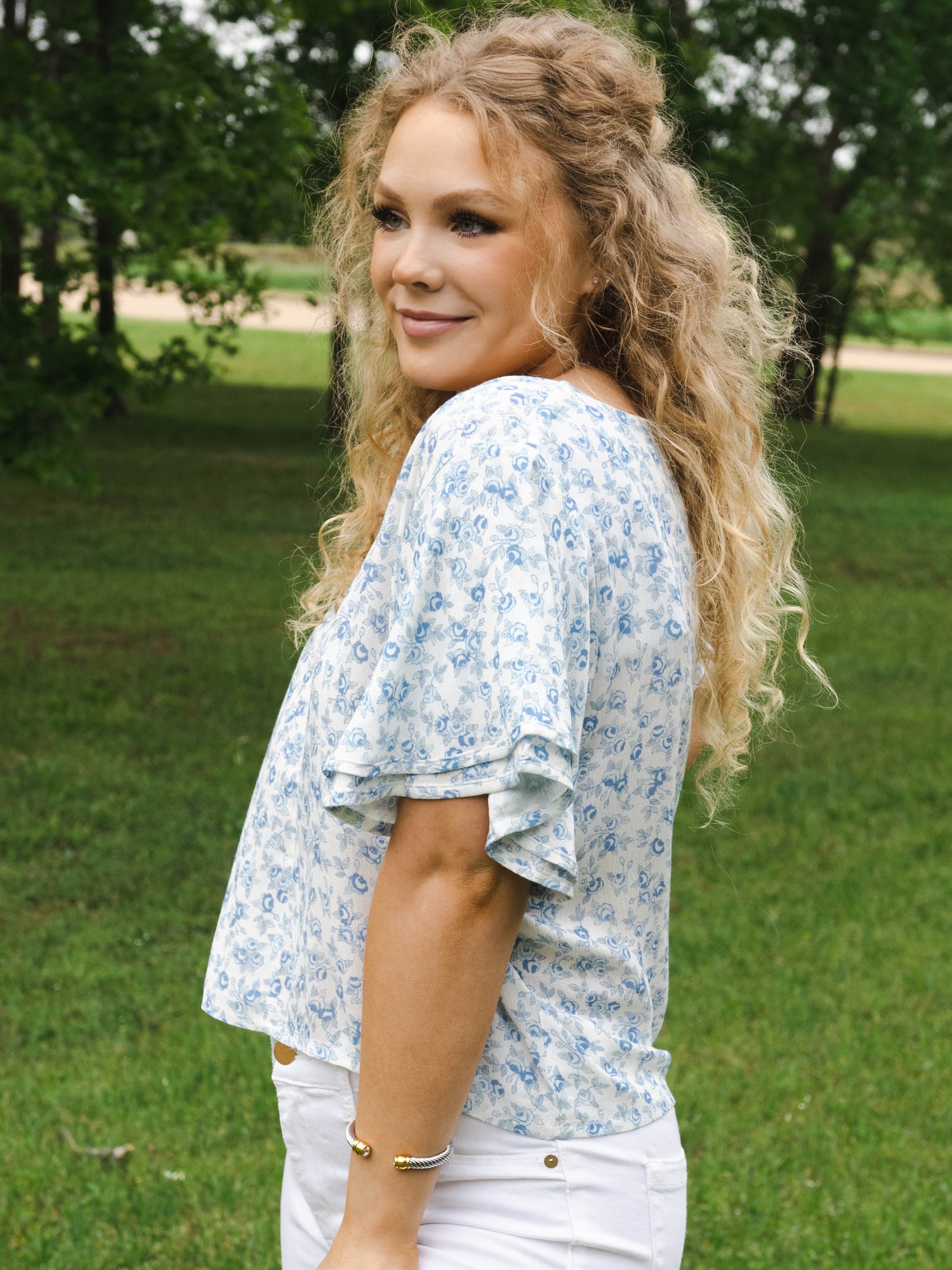 This image of a woman features the product Classic Flutter Top - Blue Jay Floral. This top has a keyhole back and flowy double ruffle sleeves. It is a blue rose pattern on a pale background.
