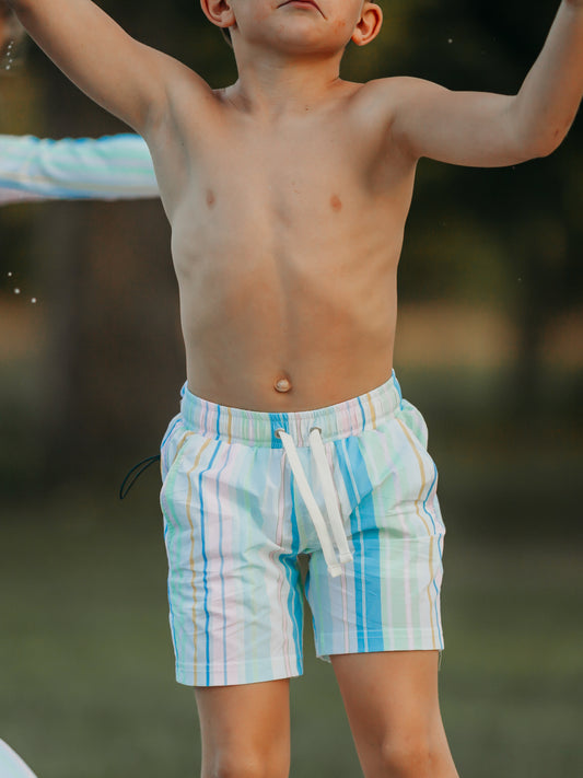 This image of a boy features the regular short style of our Boy's Everyday Lined Trunks – Retro Stripe. This pair of trunks is a pattern of blue, green, pink, orange, and white stripes.