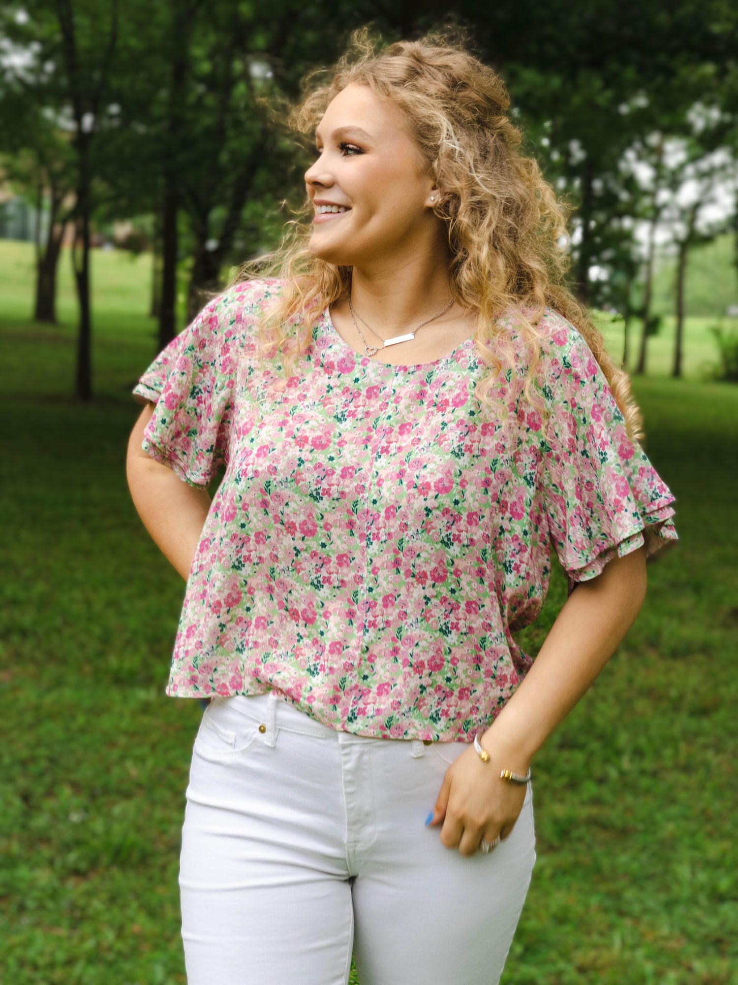 This image of a woman features the product Classic Flutter Top - Joyful. This top has a keyhole back and flowy double ruffle sleeves. It is a pink and green floral pattern.