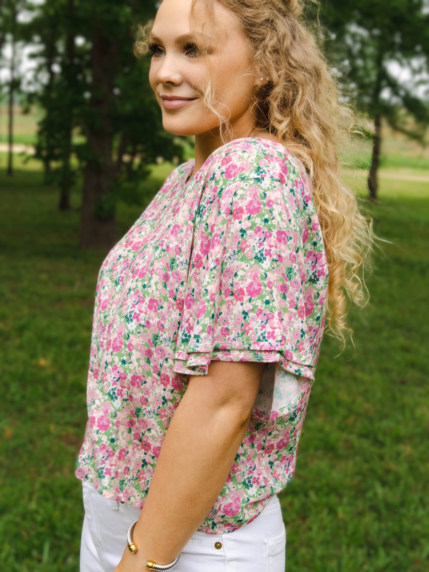 This image of a woman features the product Classic Flutter Top - Joyful. This top has a keyhole back and flowy double ruffle sleeves. It is a pink and green floral pattern.