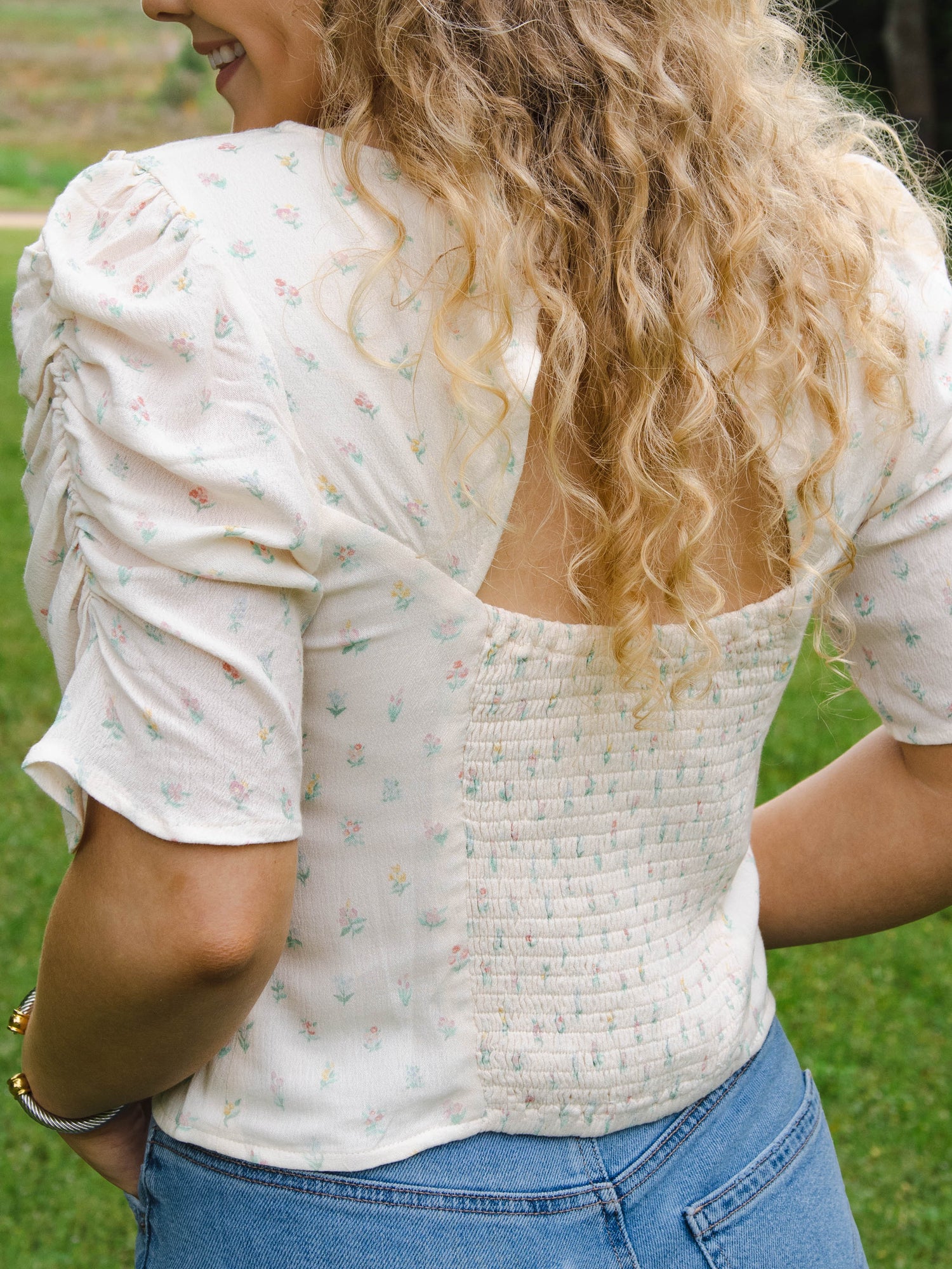 This image of a woman features the product Classic Smocked Cutout Top – Floral Drop. This short sleeve top has a smocked and keyhole back, and synched sleeves. It is a pattern of tiny hand drawn flowers spaced out across a background of cream.