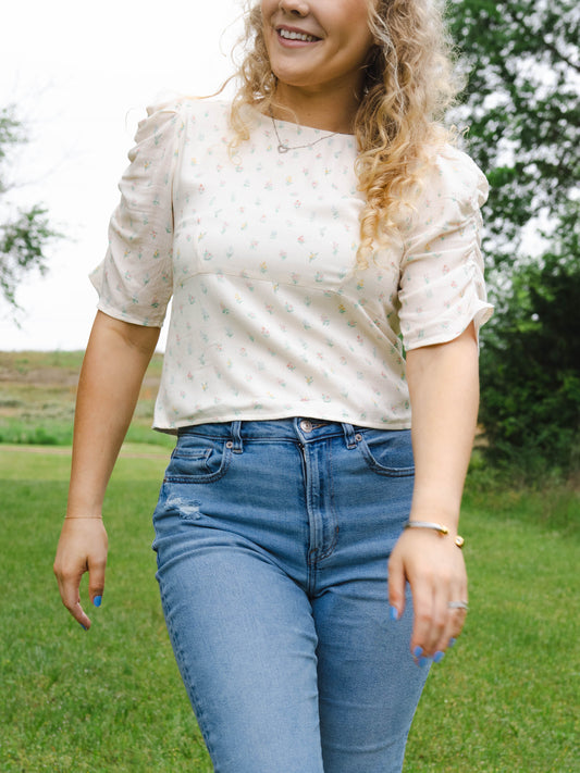 This image of a woman features the product Classic Smocked Cutout Top – Floral Drop. This short sleeve top has a smocked and keyhole back, and synched sleeves. It is a pattern of tiny hand drawn flowers spaced out across a background of cream.