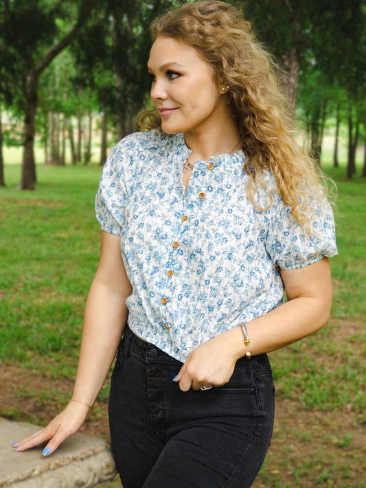This image of a women features the product Classic Button Top - Blue Jay Floral. This top has functional buttons down the bodice, puff sleeves, and elastic ruffle neckline. It is a blue rose pattern on a pale background.